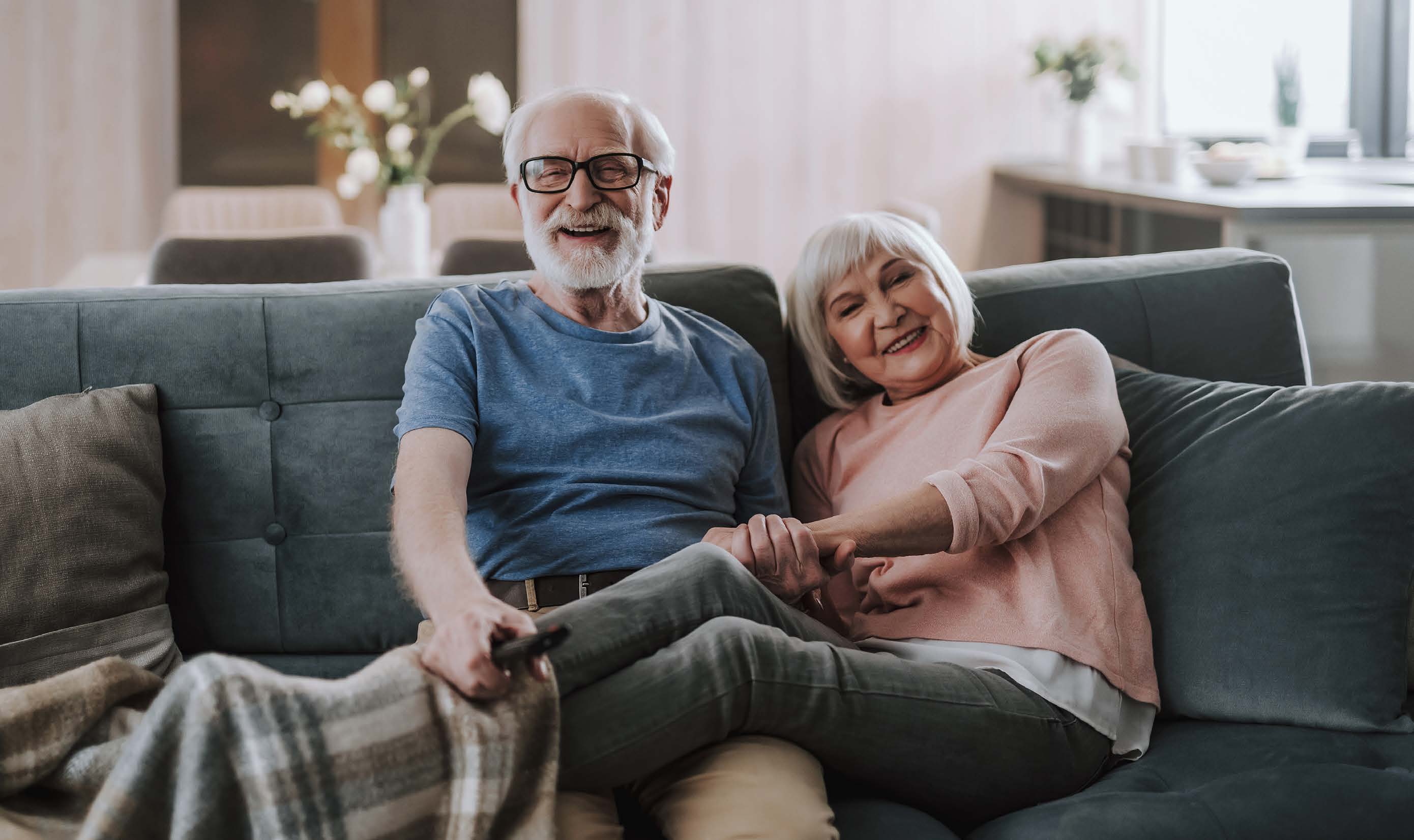 Elderly couple sitting on a couch comfortably and holding hands while laughing towards the camera