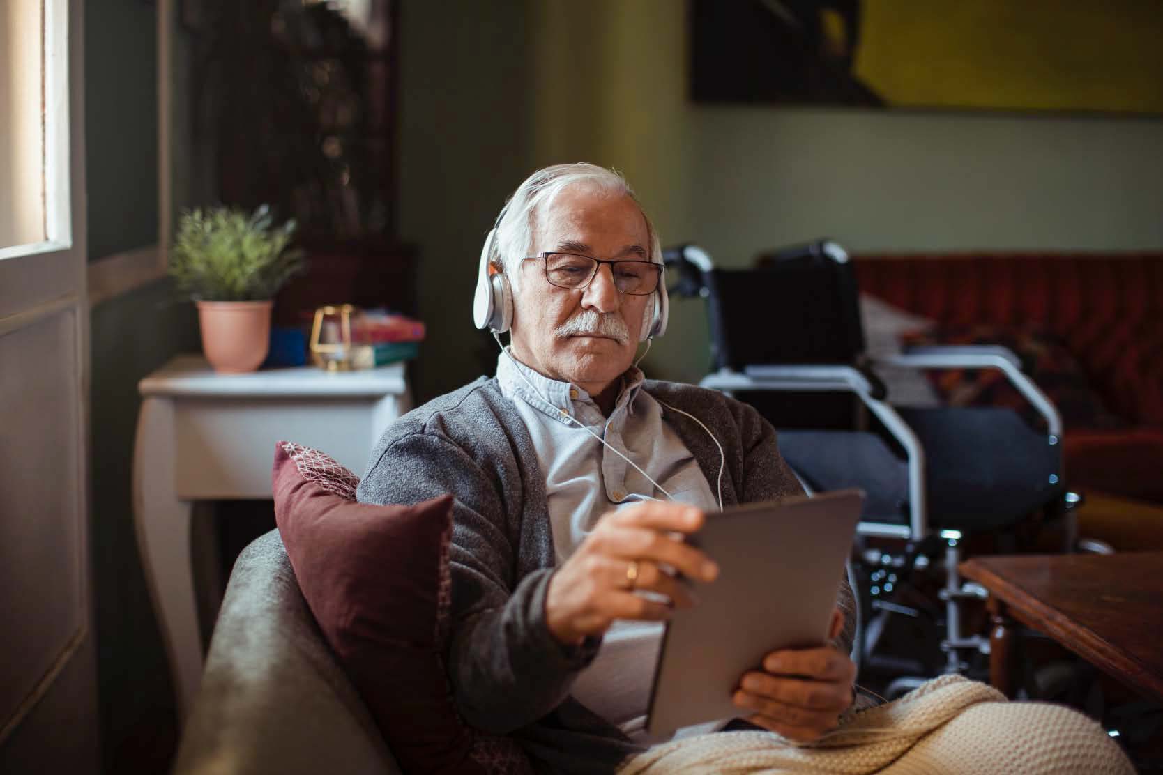 Elderly man sitting on a couch and wearing a headset while reading from his iPad